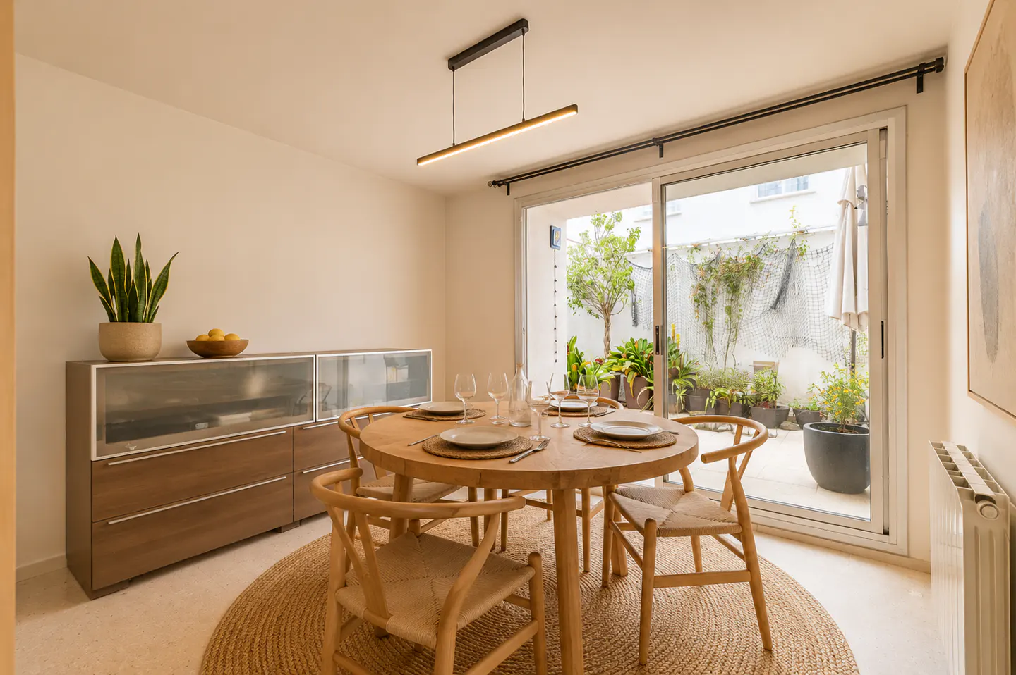 Bright dining room with a round wooden table set for five, surrounded by woven chairs on a round rug. Sliding glass doors open to a patio with plants.