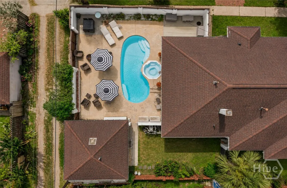 Aerial view of a backyard with a kidney-shaped pool, hot tub, two striped umbrellas, lounge chairs, and a brown roof house.