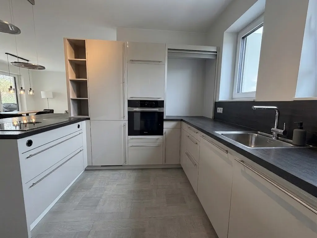 A modern kitchen with white cabinets, a black countertop, and stainless steel sink. A built-in oven and island are also visible.