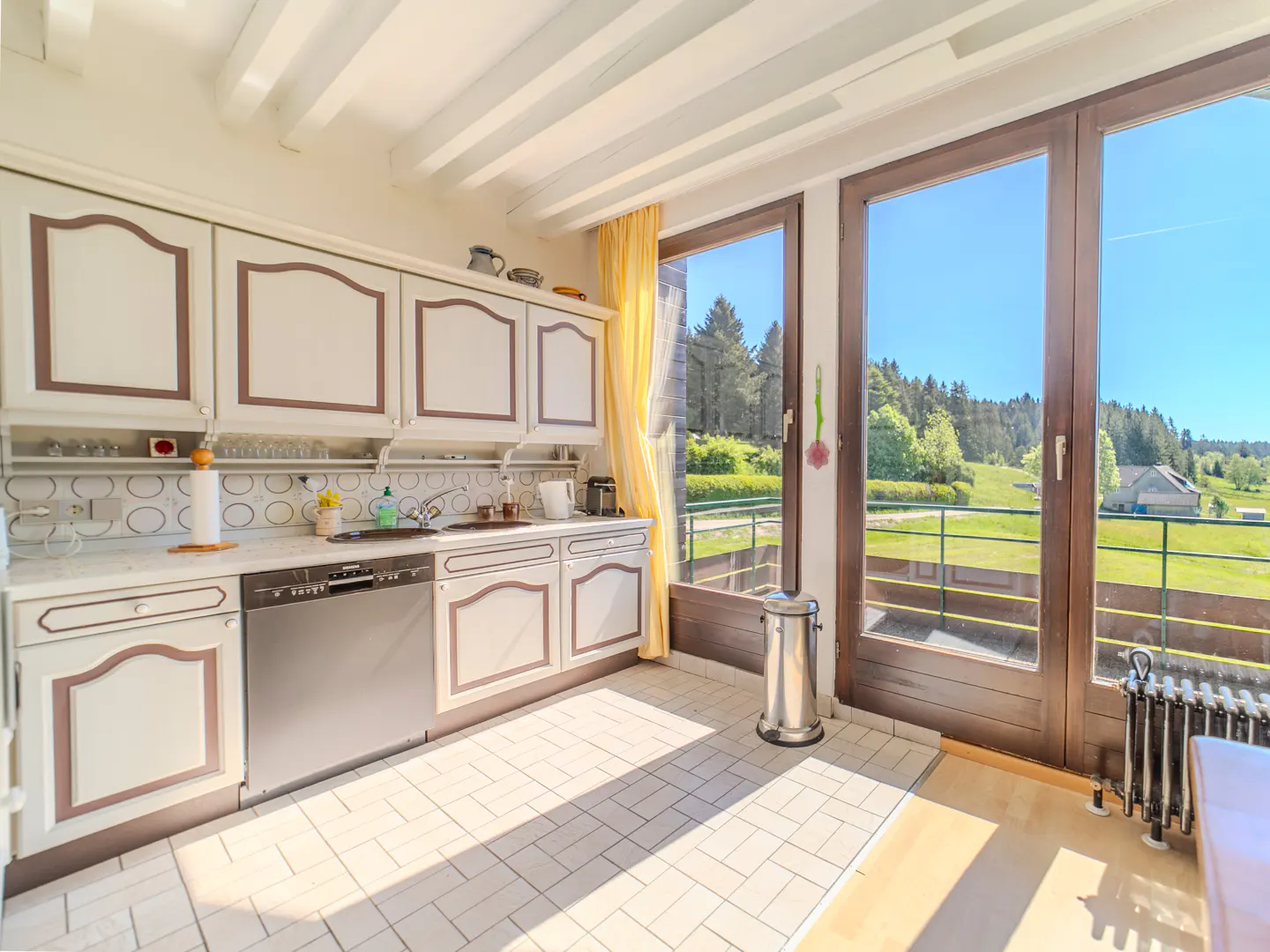 Bright kitchen with white cabinets, brown trim, and large windows overlooking a green landscape.