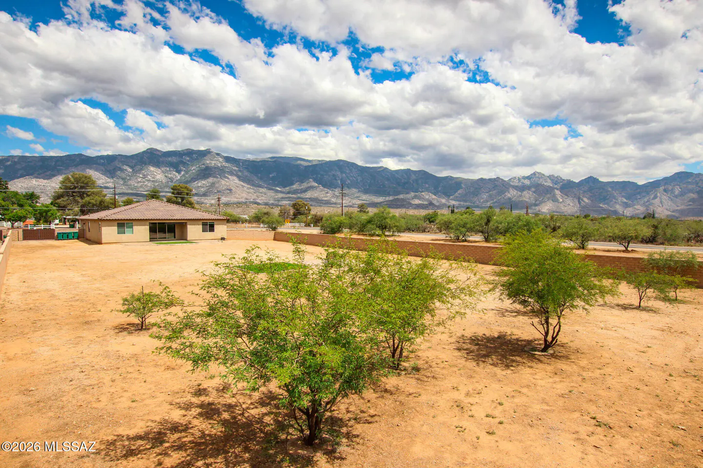 A tan house sits on a large, dry lot with trees. Mountains are in the background under a blue sky with white clouds.