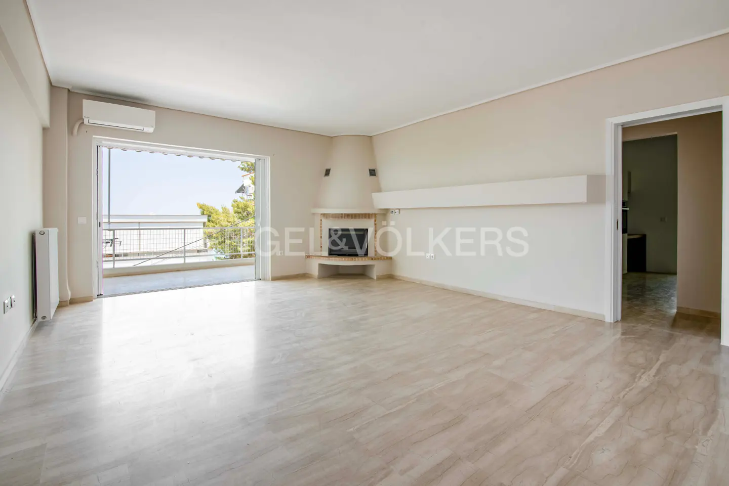Bright, empty living room with marble floors, a fireplace, and a balcony with a view. Cream-colored walls and a doorway to another room.