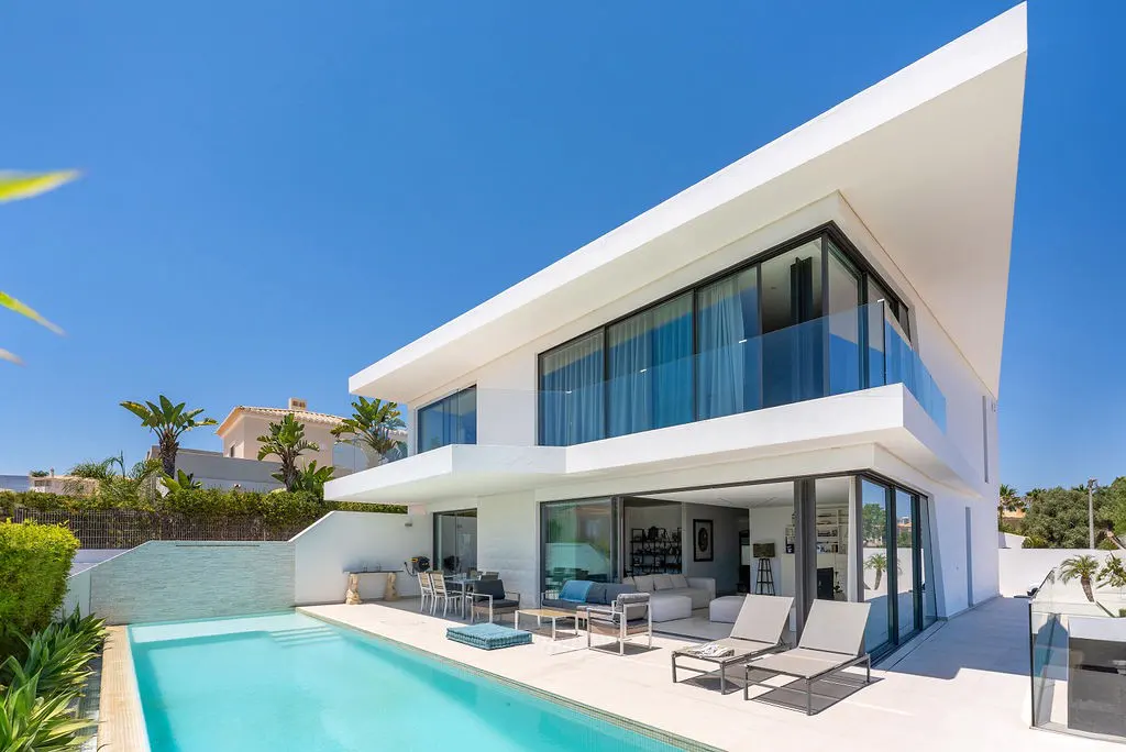 Modern white two-story house with a pool. Lounge chairs sit on the patio, with a view of the open living room. Blue sky.