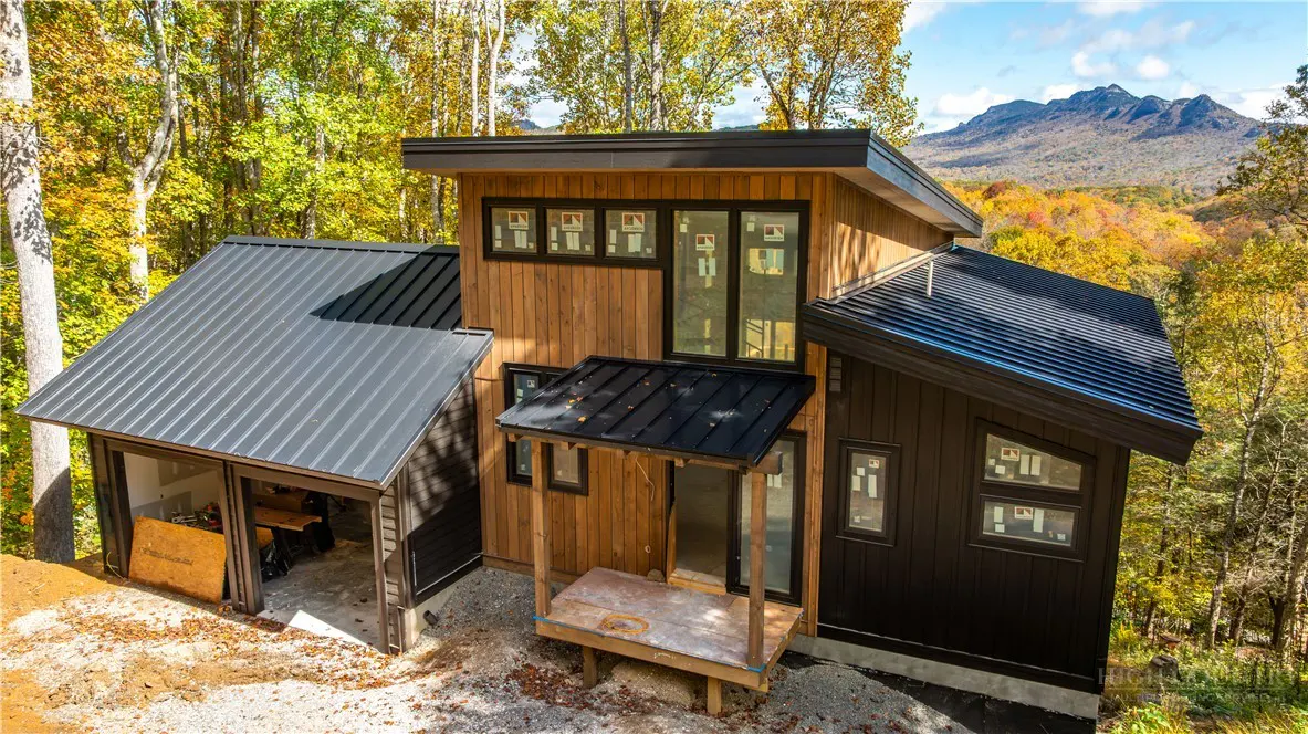 Modern wood home with black trim and metal roof, nestled in a forest with fall foliage and mountain views.