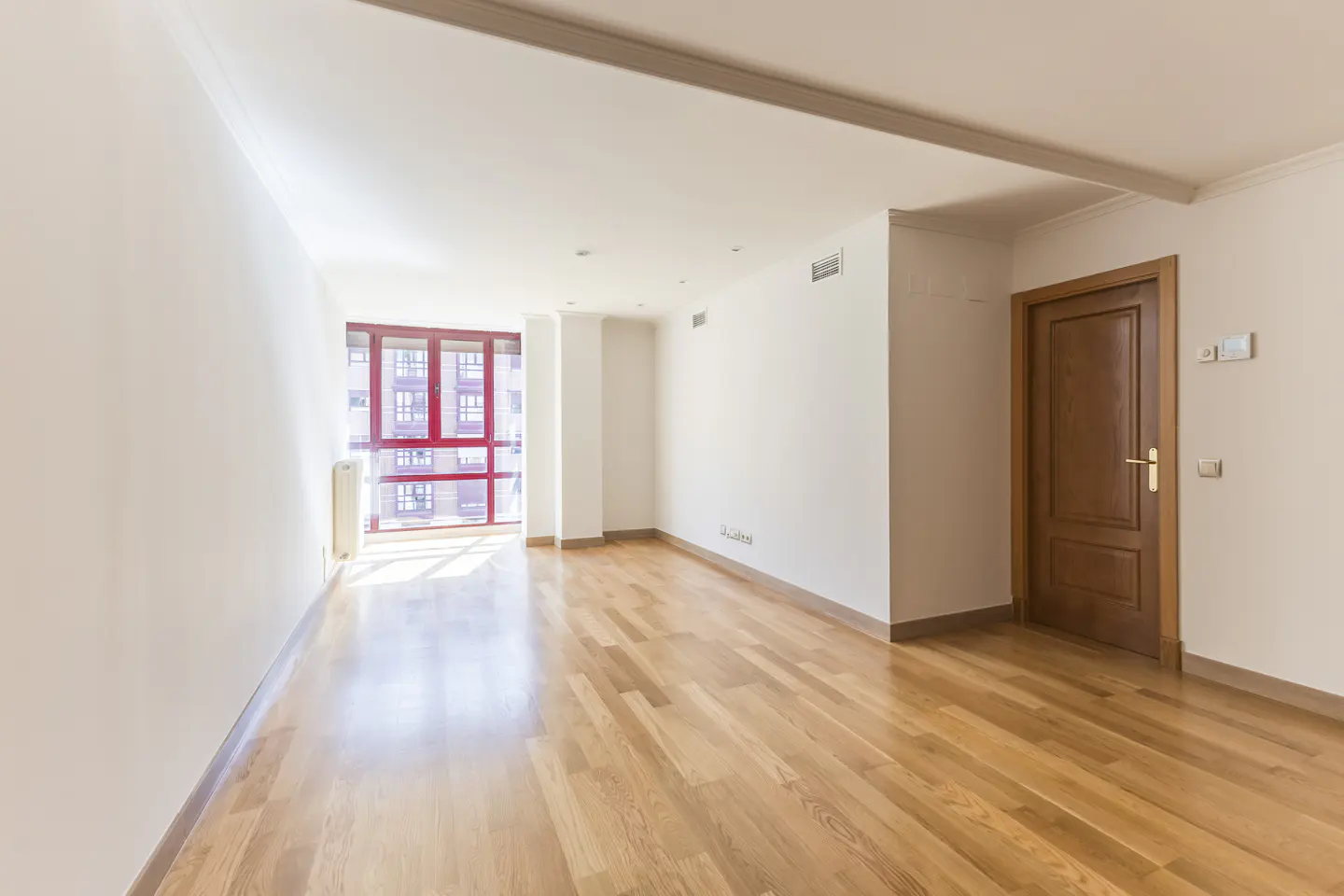 Bright, empty room with hardwood floors, white walls, and a red-framed window. A wooden door is on the right.