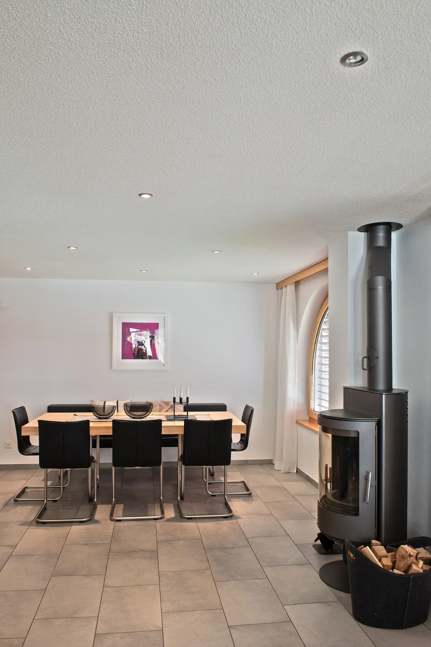 A modern dining room with a wood-burning stove. The room has a long table with black chairs, a white wall with art, and a tiled floor.