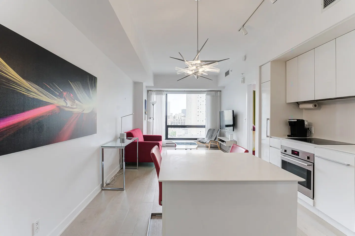 Bright, modern condo interior with white walls, light wood floors, and red accents. Kitchen island in foreground, living area with city view in background.