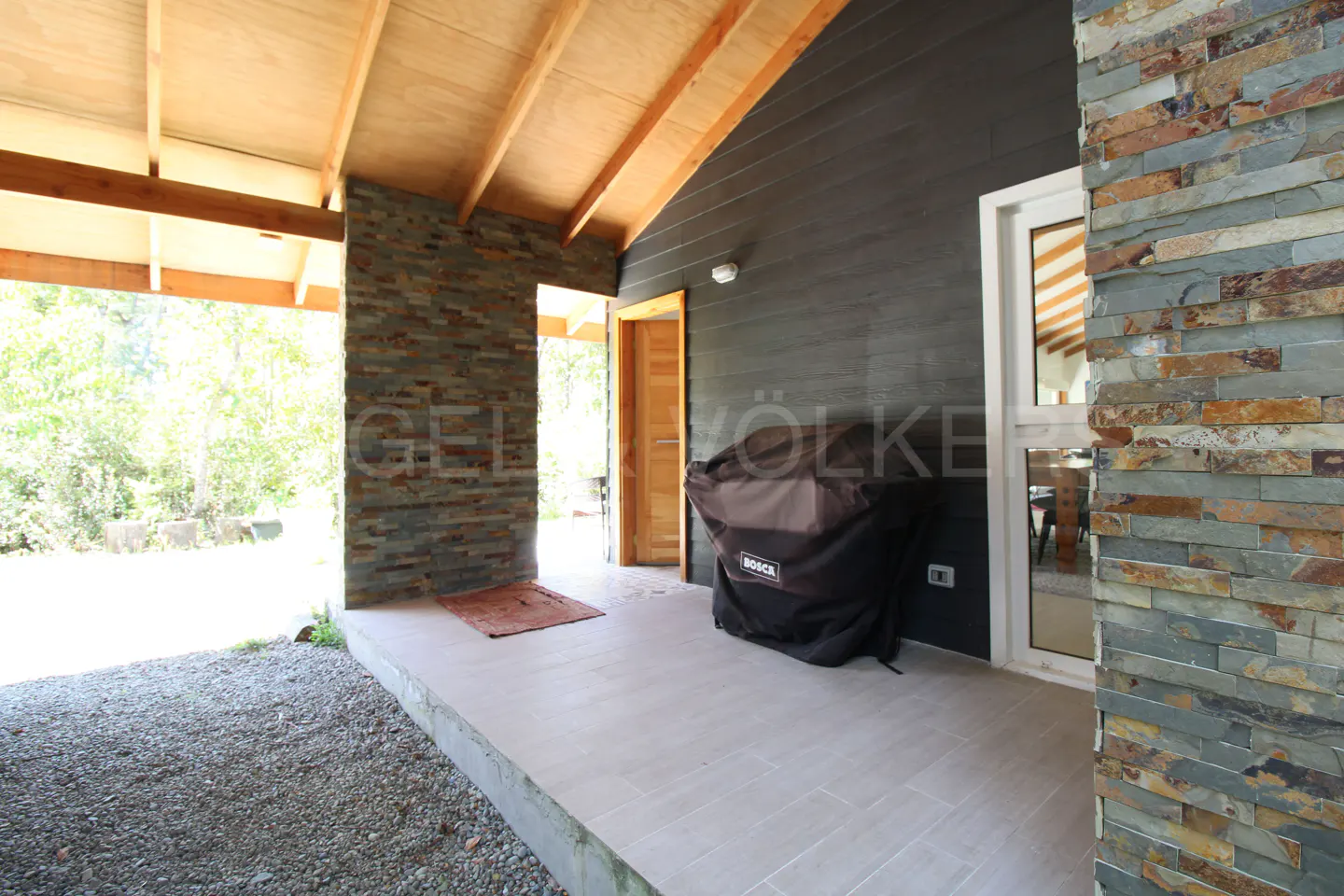Covered patio with stone accent wall, wood ceiling, and black siding. A grill with a black cover sits against the wall.