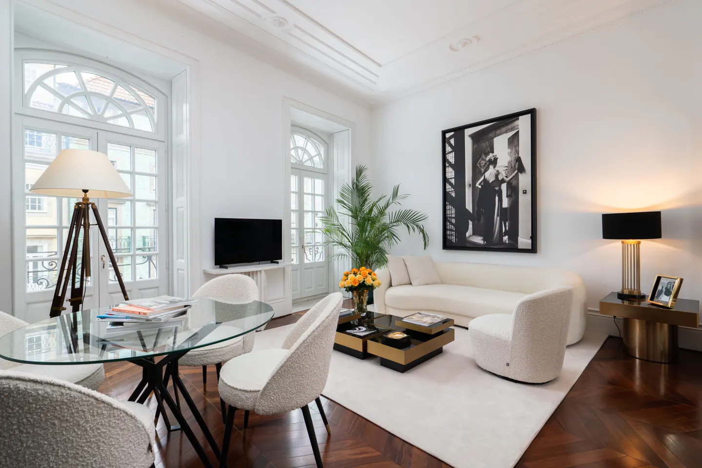 Bright living room with white walls, herringbone wood floors, and large windows. A glass table with white chairs sits near a tripod lamp. A white sofa and black and gold coffee table are on a white rug.