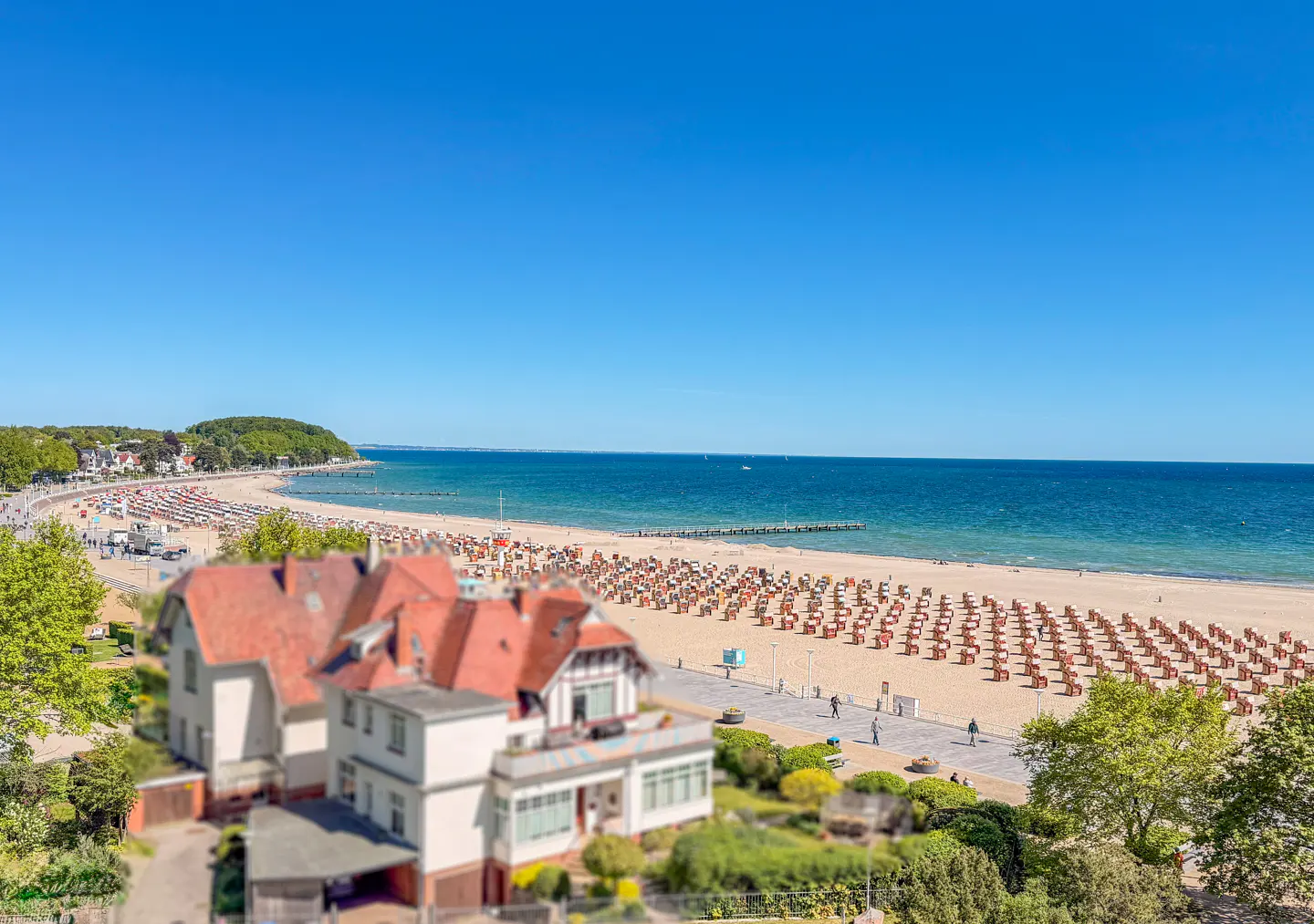 Beach view with rows of beach chairs, blue sea, and a building with a red roof in the foreground on a sunny day.