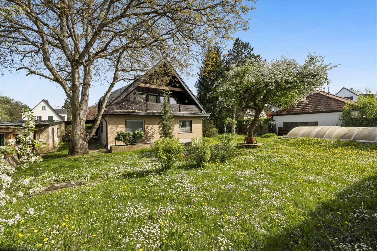 A tan house with a dark roof sits on a green lawn dotted with white and yellow flowers. Trees surround the house under a blue sky.