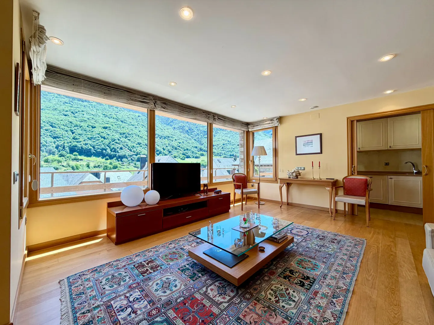 Living room with large windows, mountain views, a glass coffee table, patterned rug, and an open doorway to the kitchen.