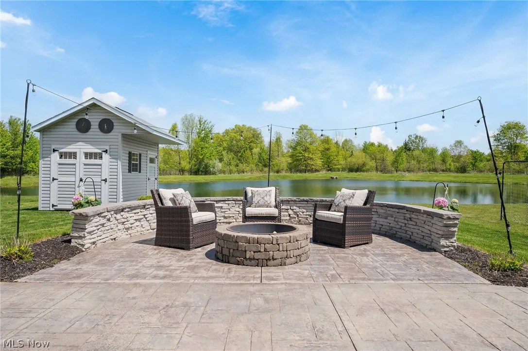 Outdoor patio with a stone fire pit, three wicker chairs with cushions, and string lights. A pond and shed are in the background.