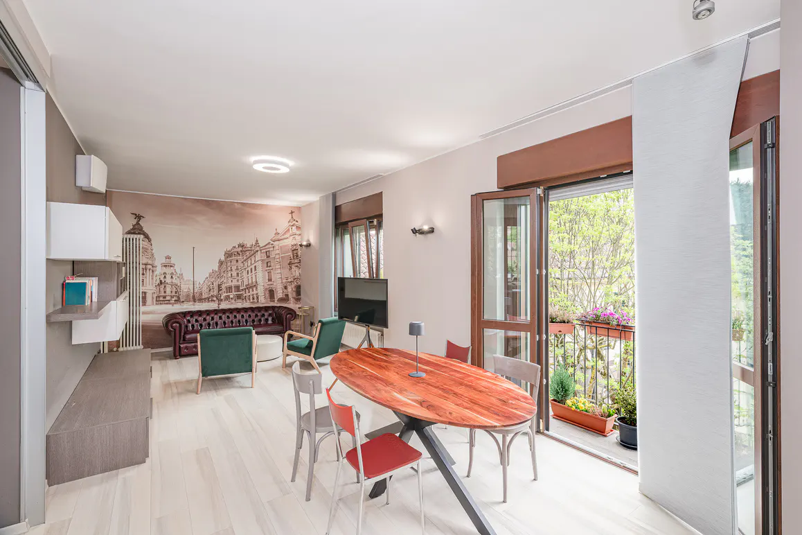 Bright living room with wood floors, oval table, and balcony. Cityscape mural behind a brown leather sofa and green chairs.