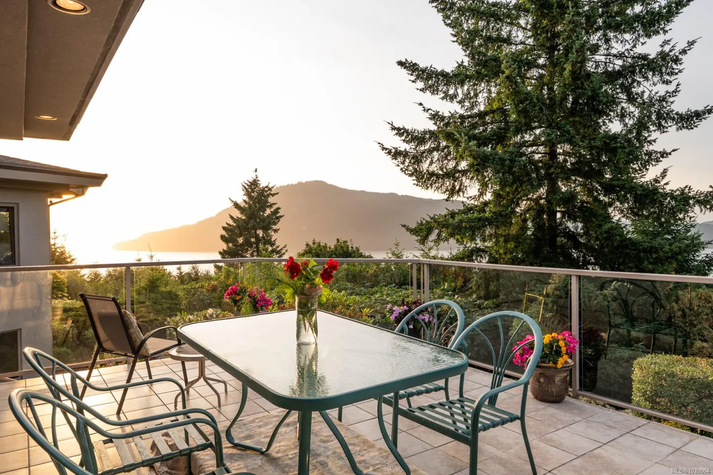 Patio with glass table, chairs, and flower vase overlooking a scenic sunset view of mountains and water.