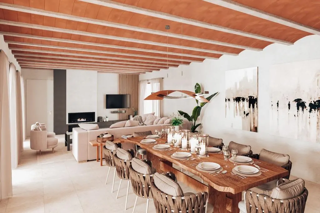 A bright, open-plan living and dining area with a long wooden table set for a meal, surrounded by woven chairs. A sofa, fireplace, and TV are in the background.