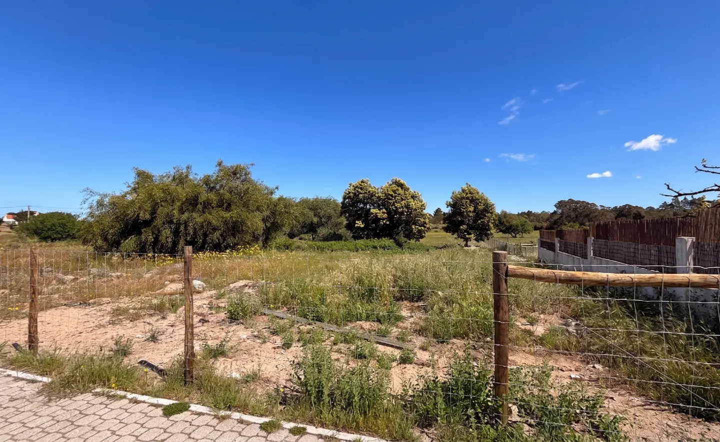 Vacant lot with sandy soil, tall grass, and trees under a blue sky. A wire fence with wooden posts surrounds the property.