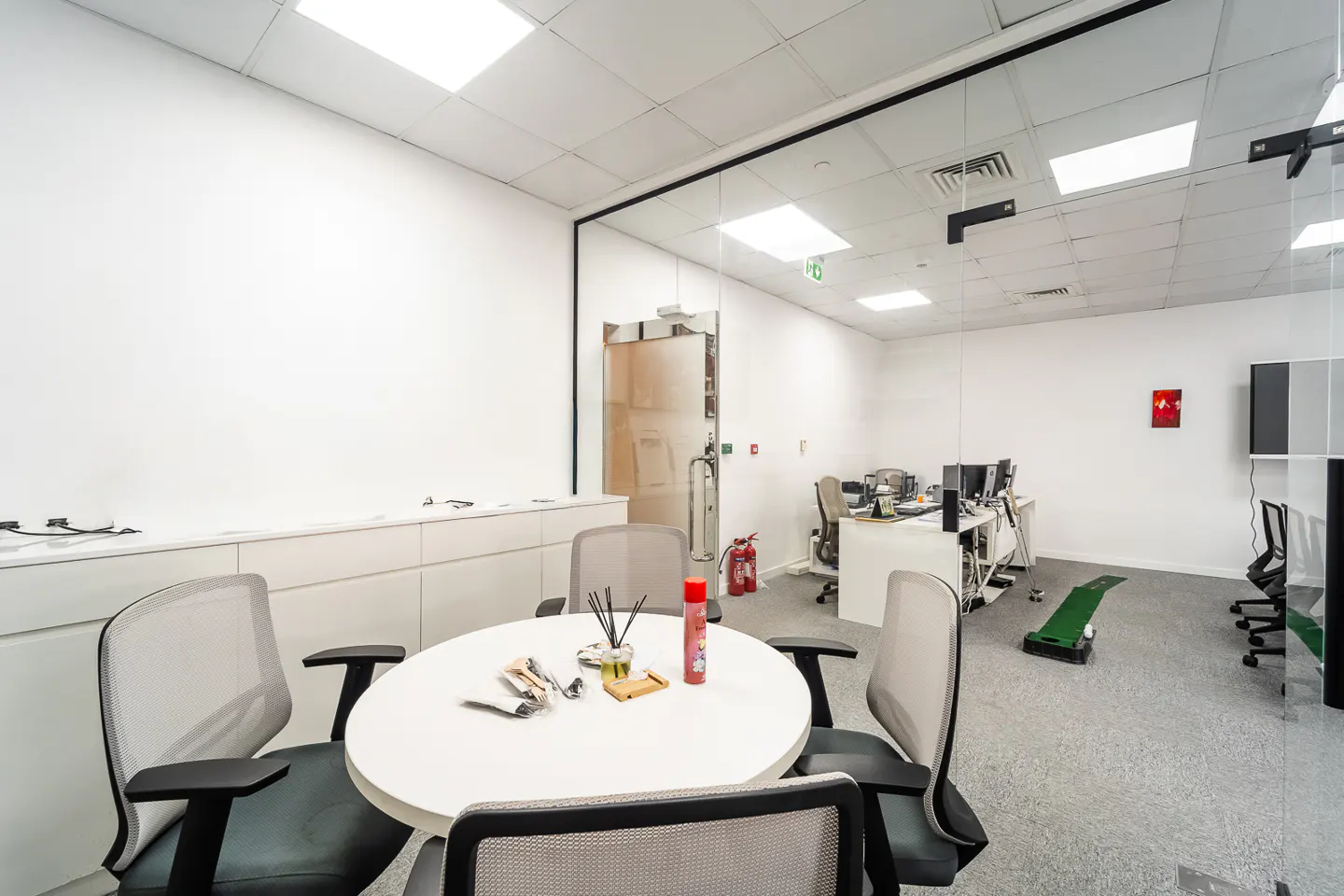 Bright office space with a white round table surrounded by four chairs in the foreground. Desks and computers are visible in the background.