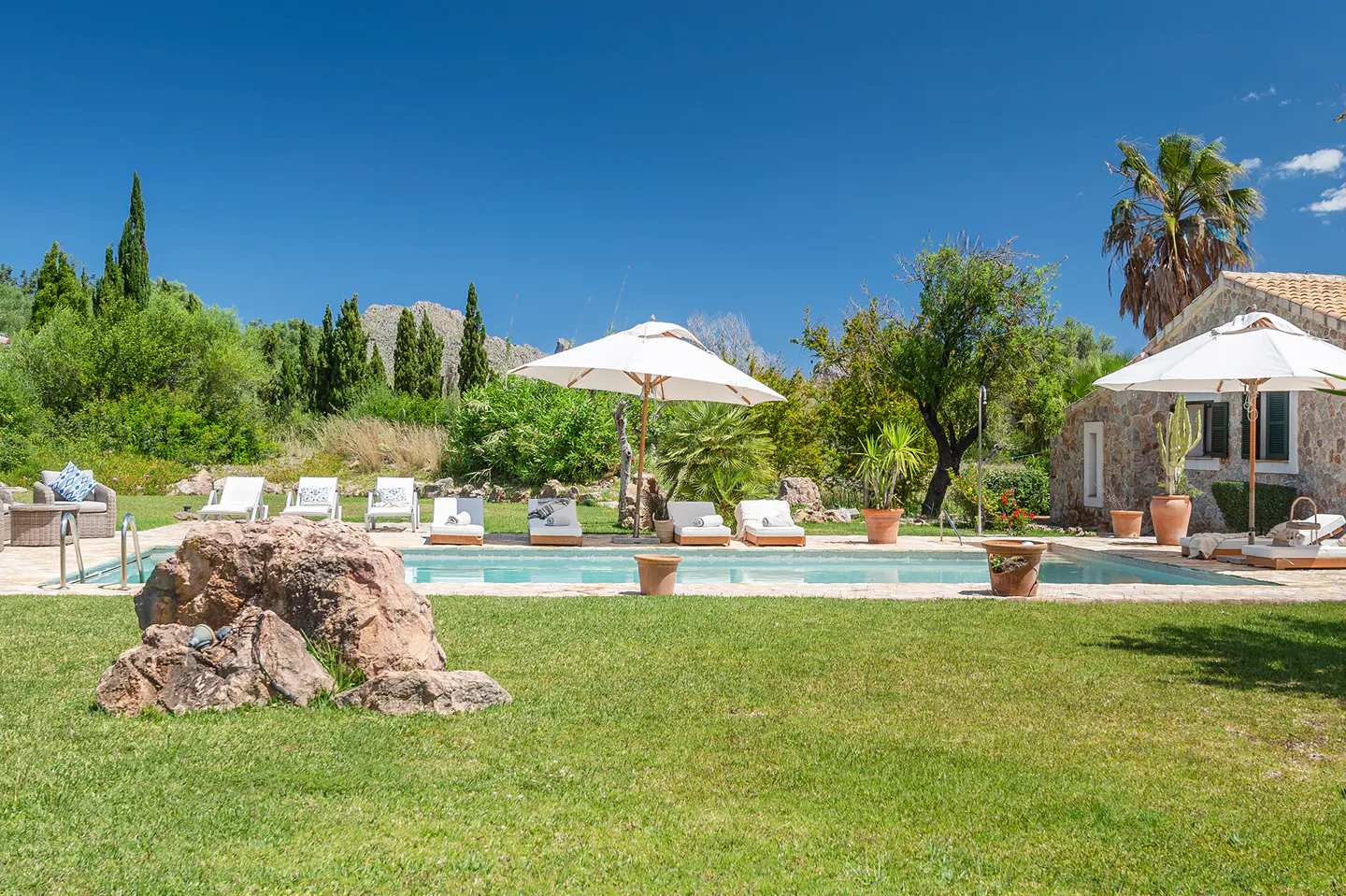 Outdoor pool with white lounge chairs and umbrellas on a green lawn, with a stone house and trees in the background.