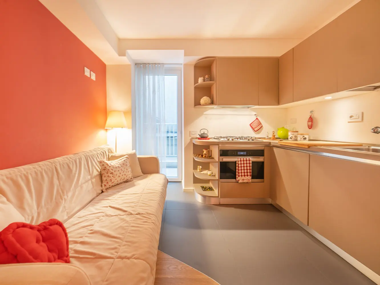 Cozy apartment interior with a beige sofa, red heart pillow, and a modern kitchen with tan cabinets. A balcony door lets in natural light.