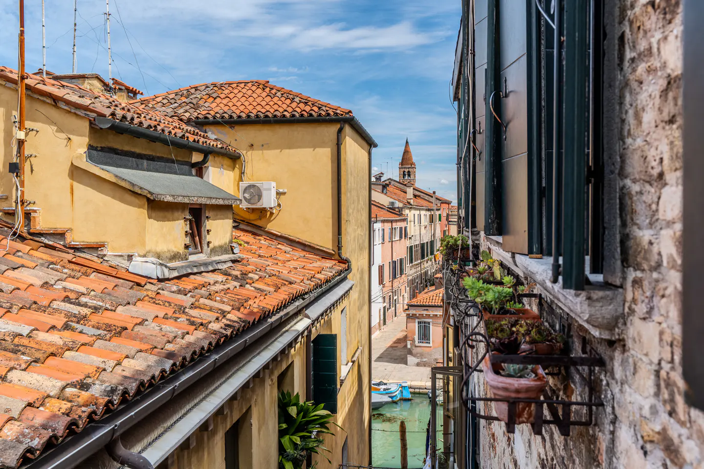 View of a Venice canal from a window with plants. Buildings with orange tile roofs line the canal under a blue sky.
