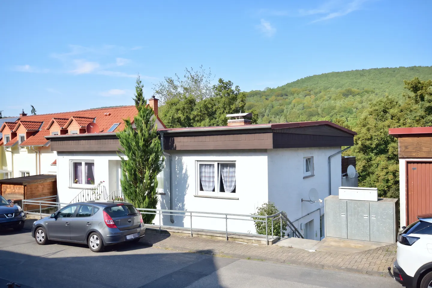A white, single-story house with a brown roof, a gray car parked in front, and a wooded hill in the background.