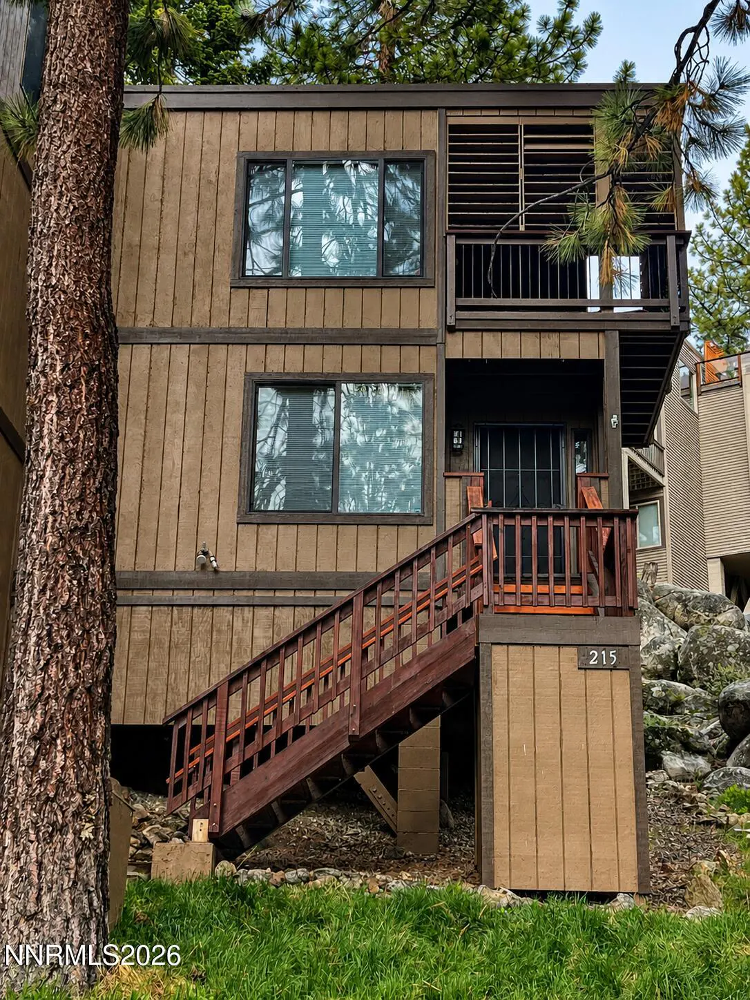Exterior view of a brown, two-story condo with a wooden staircase leading to a deck. Large tree on the left. Number 215 visible.