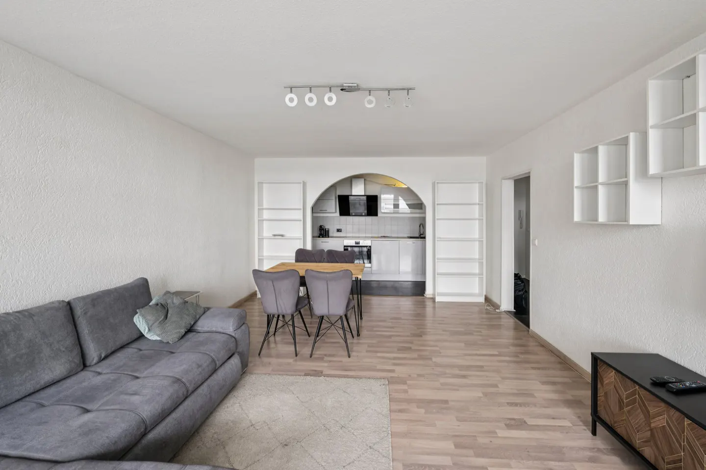 A bright, open-concept living space with a gray sofa, dining table, and kitchen visible through an arched opening. White walls and light wood floors create a modern feel.