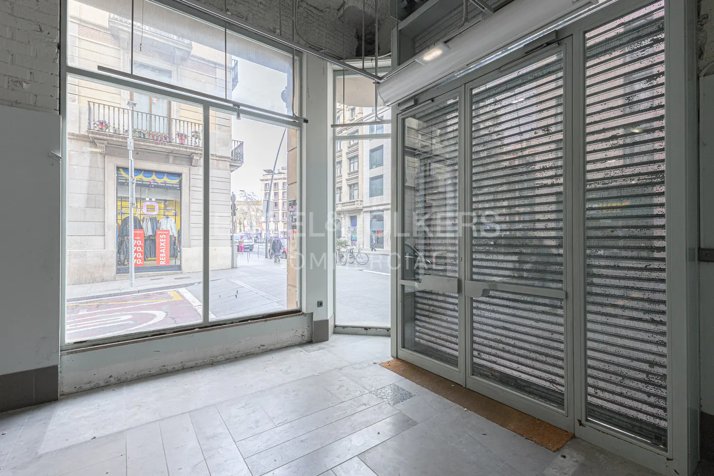 Empty commercial space with large windows and metal security shutters. Street view visible through the windows. Interior is gray and white.