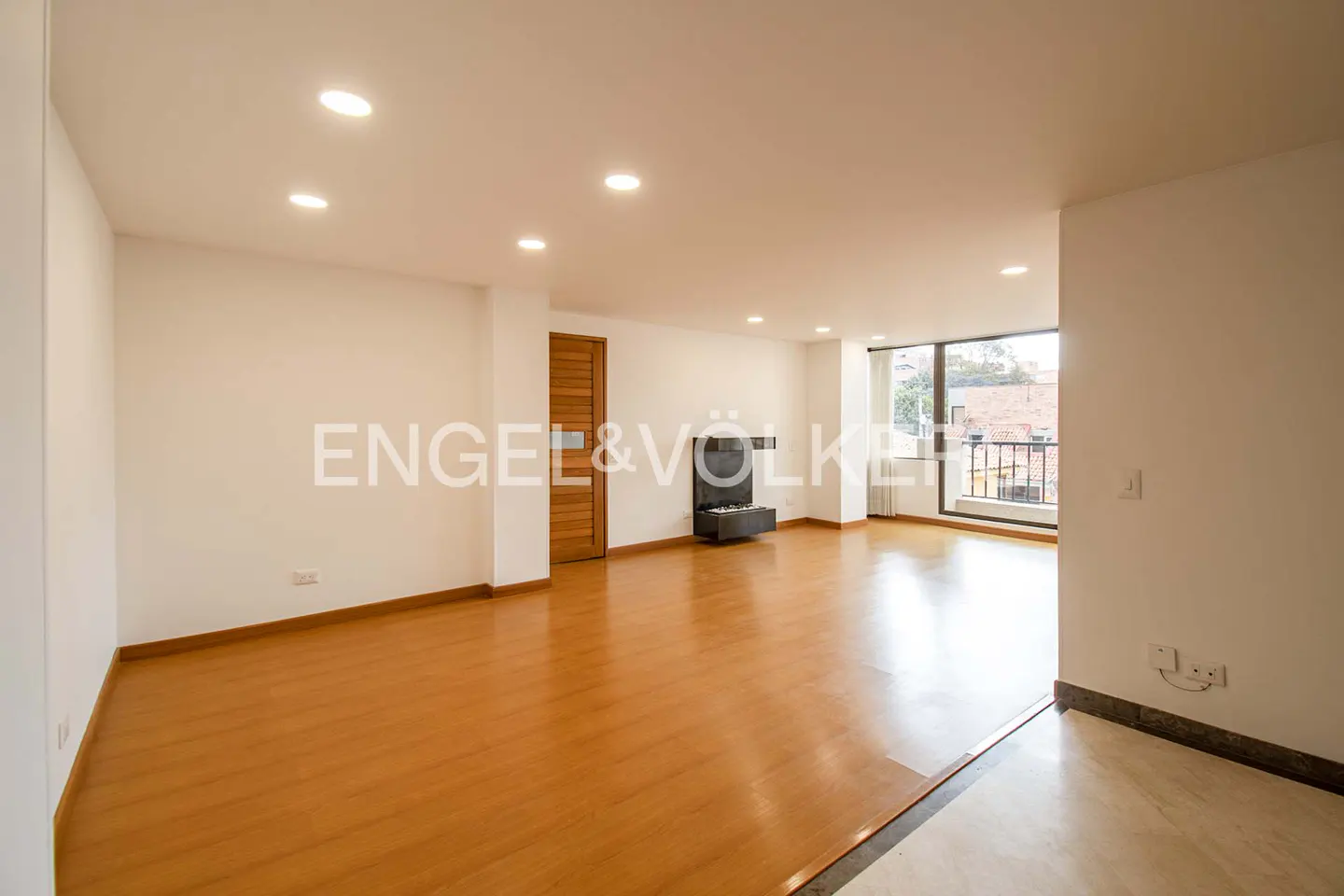 Bright, empty living room with hardwood floors, white walls, and a modern fireplace. Balcony with city view. Engel & Völkers logo visible.