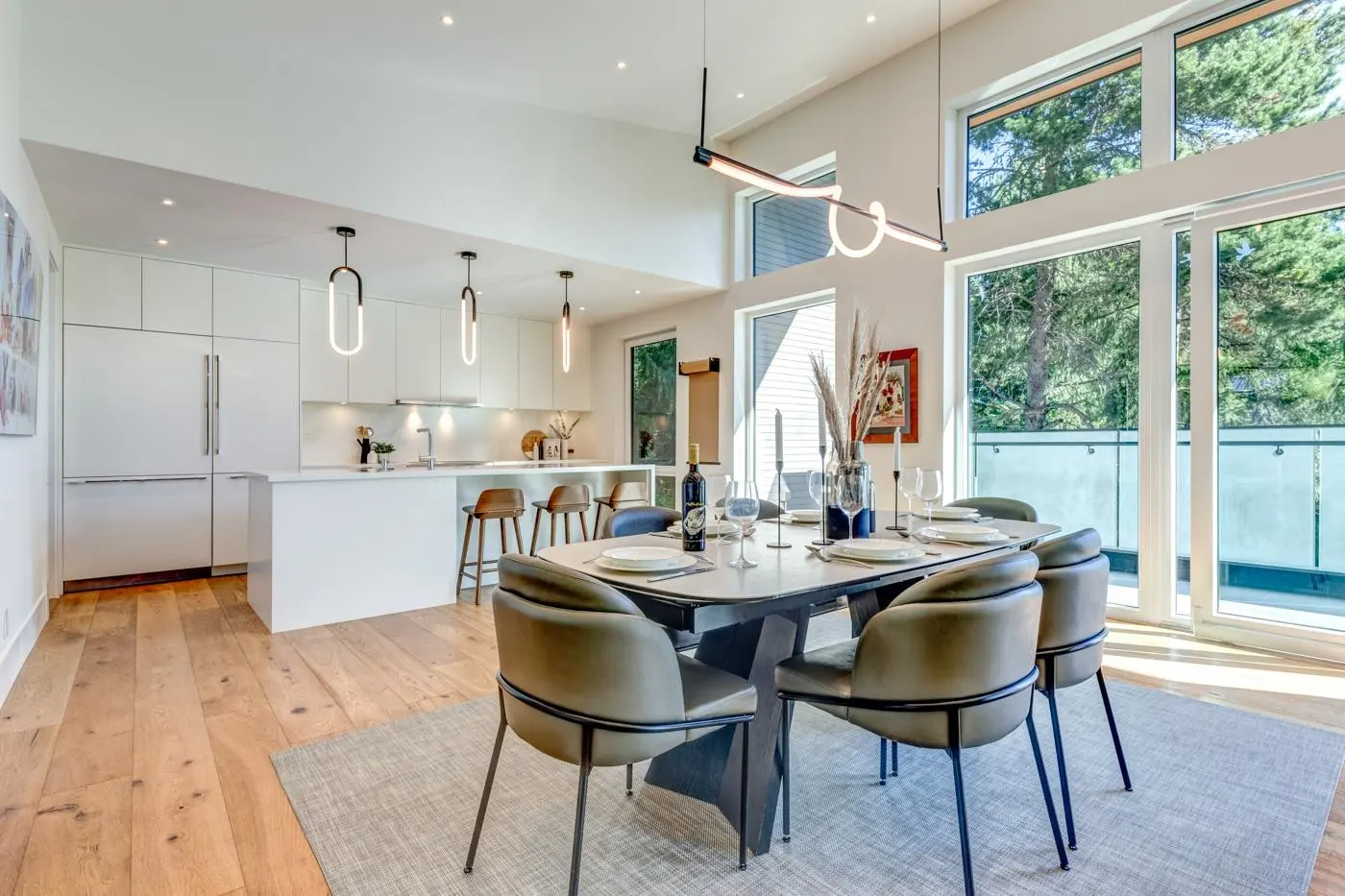 Bright, open-concept kitchen and dining area with white cabinets, wood floors, and a dining table set for a meal. Large windows offer a view of trees.