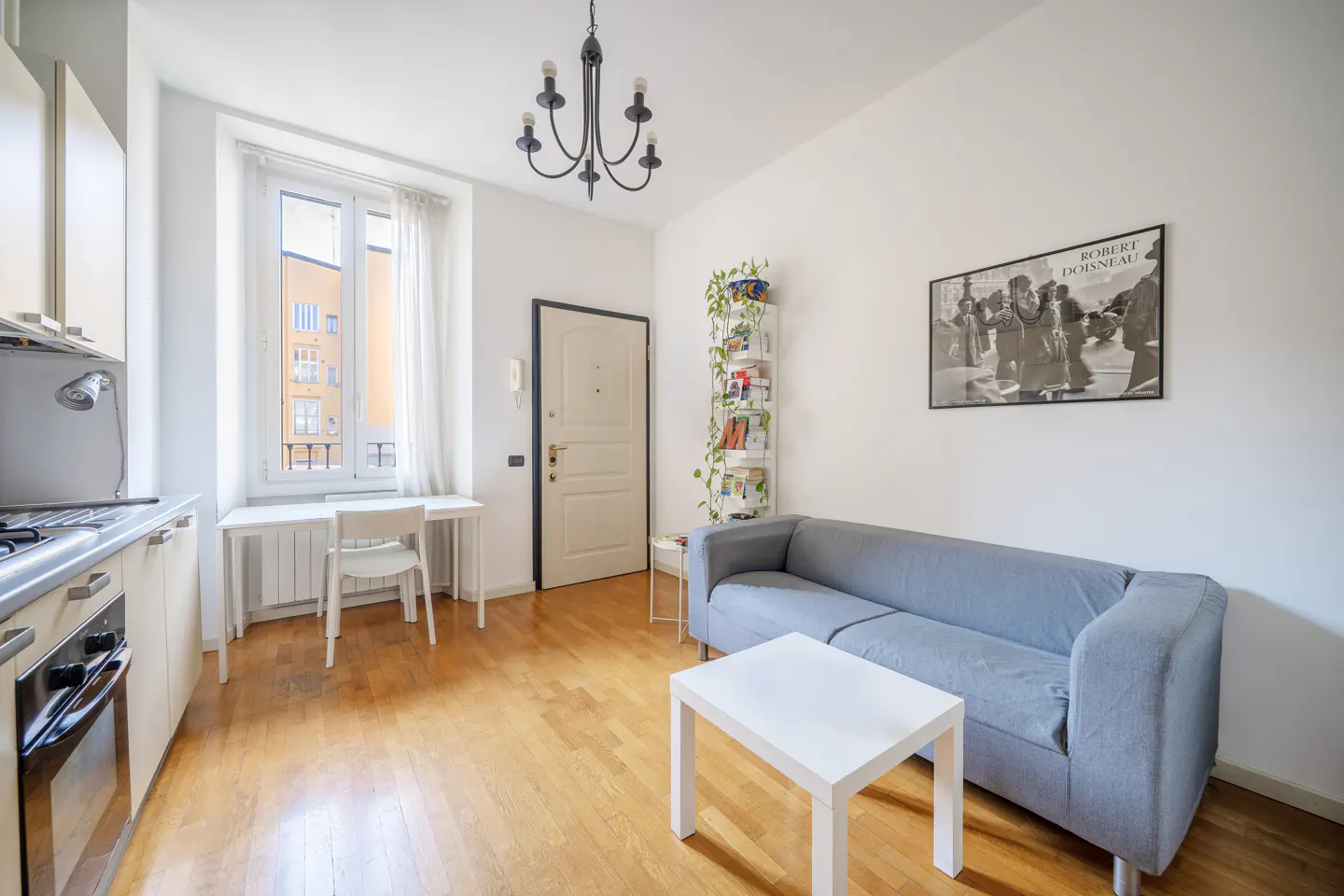 Bright apartment interior with hardwood floors, white walls, and a gray sofa. A black chandelier hangs above a white coffee table. Kitchen area visible.