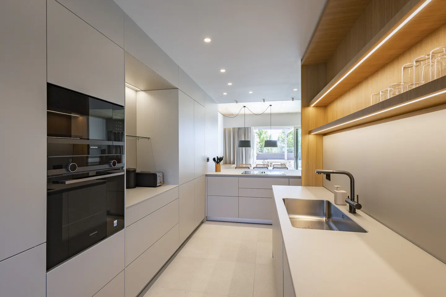 Bright, modern kitchen with white cabinets, stainless steel sink, and black faucet. Wood shelves with glasses and recessed lighting add warmth.