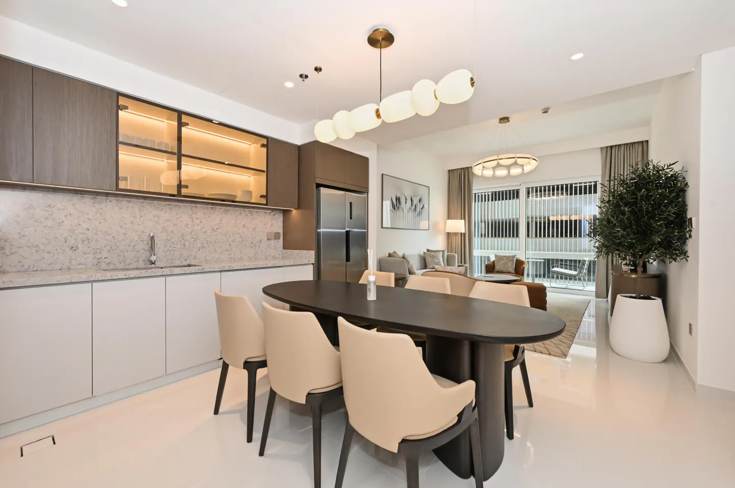 Bright, modern apartment interior with a dark oval dining table, beige chairs, and a kitchen with white cabinets. A living room is visible in the background.