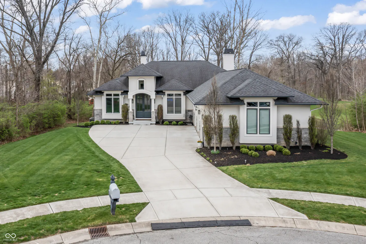 A modern, single-story home with a gray roof, white exterior, and a long concrete driveway. Green lawn and trees surround the house.