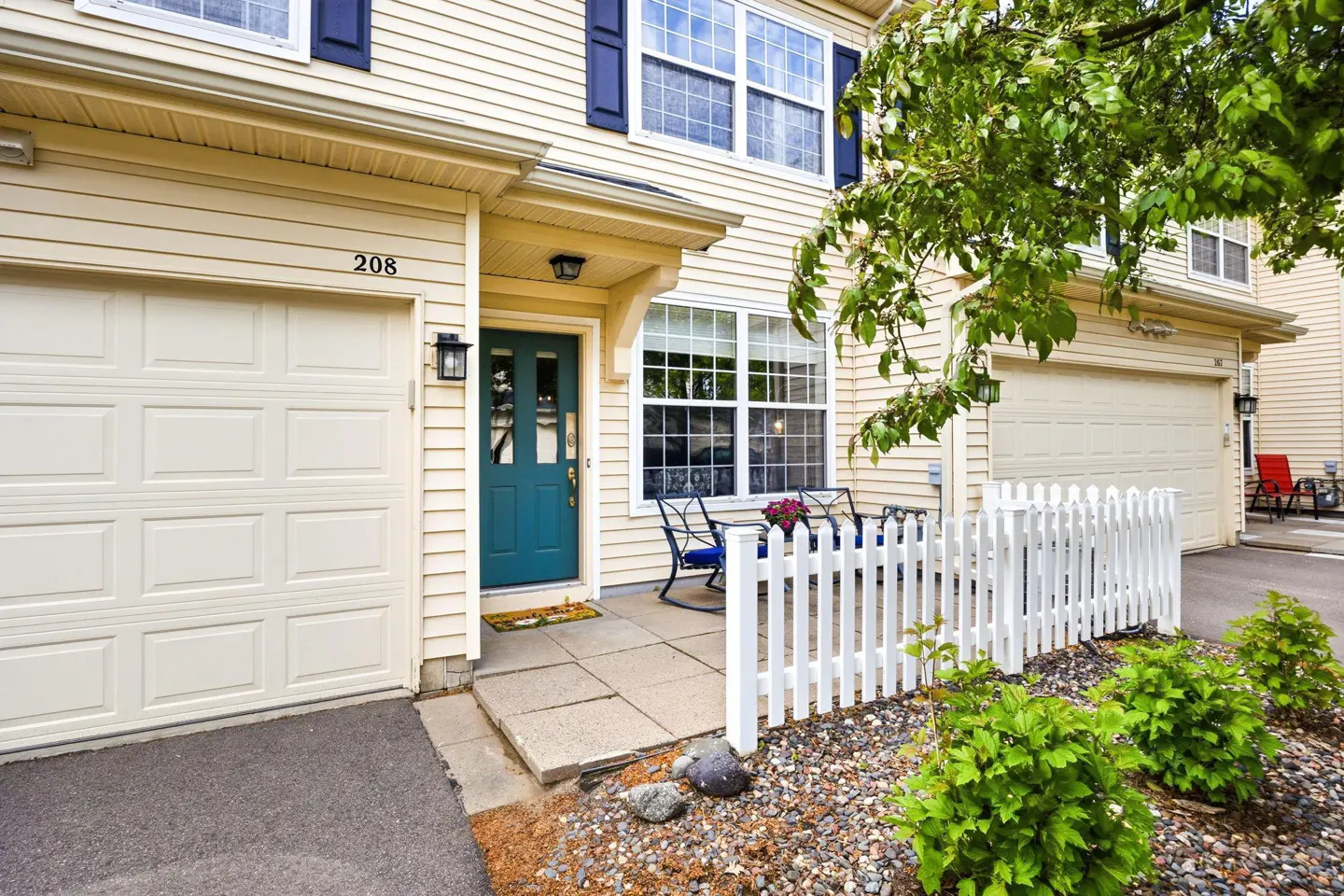 Beige two-story townhouse with a teal door, white garage door, and white picket fence. A small patio with blue chairs is visible.