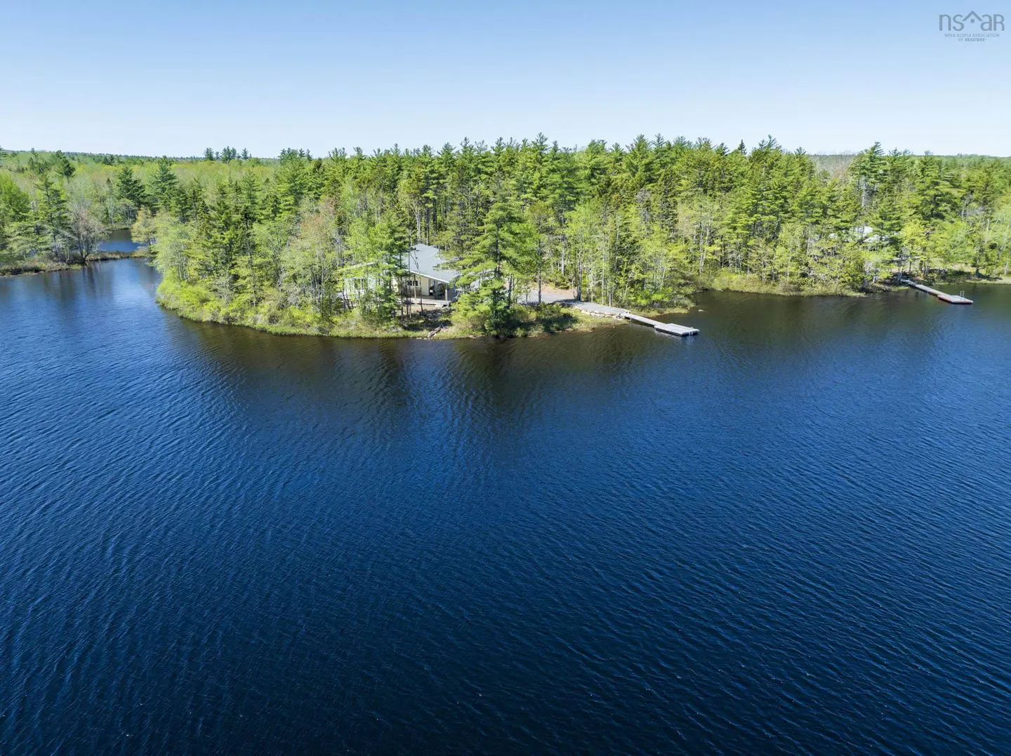 Aerial view of a lake house on a wooded peninsula with a dock, surrounded by dark blue water.