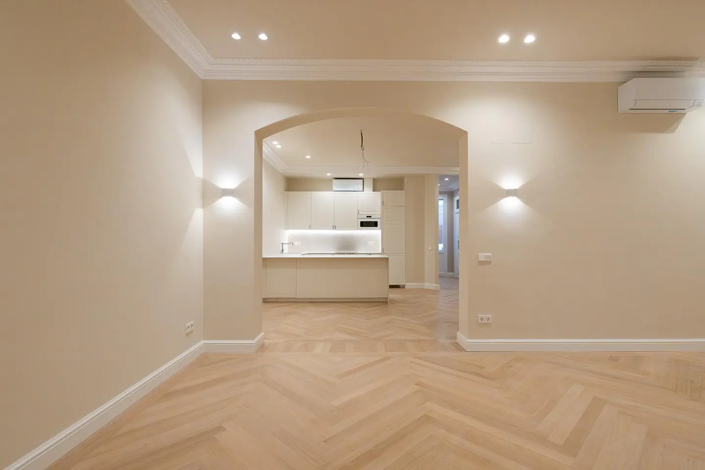 Interior view of a beige room with herringbone wood floors, an arched doorway to a white kitchen, and modern wall sconces.