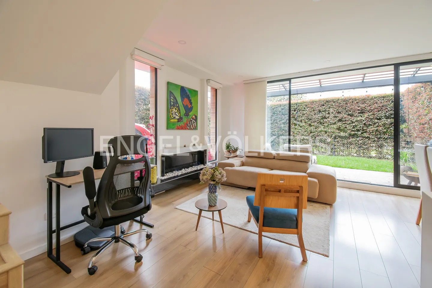 Bright living room with desk, fireplace, and tan sofa. Large windows overlook a green hedge. Wood floors and white walls.