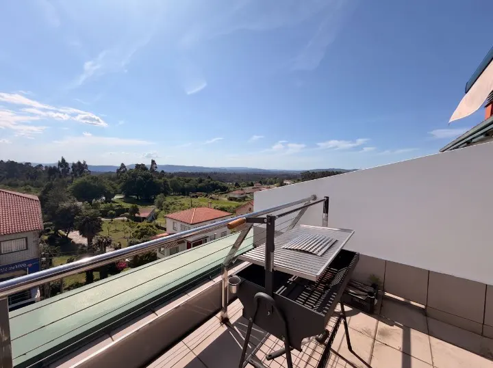 Balcony view with a black grill, metal railing, and a scenic backdrop of green trees and houses under a blue sky.