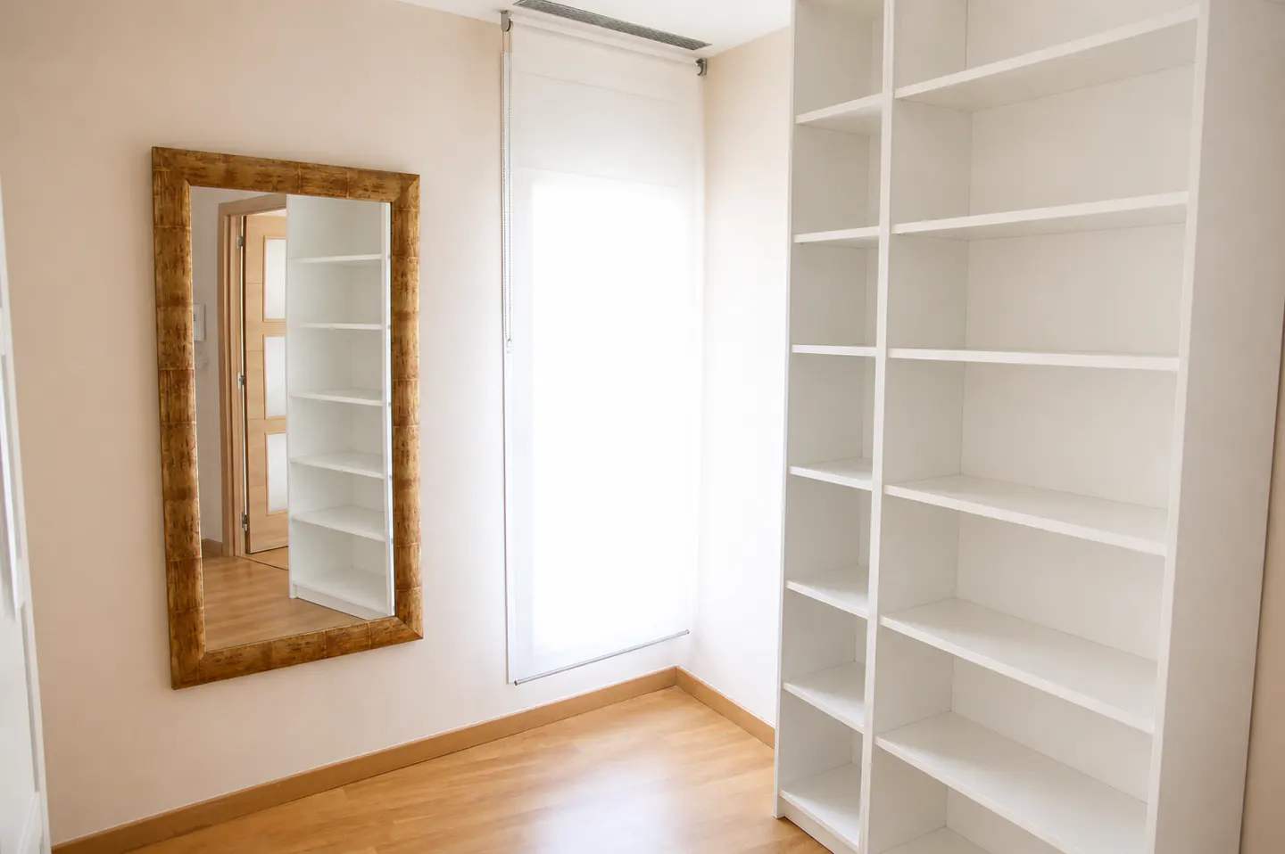 A bright room with a wood floor, a gold-framed mirror, a white window shade, and a large white bookcase.