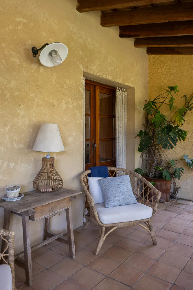 Outdoor patio with a wicker chair, blue pillows, a wooden table with a lamp, and a potted plant.