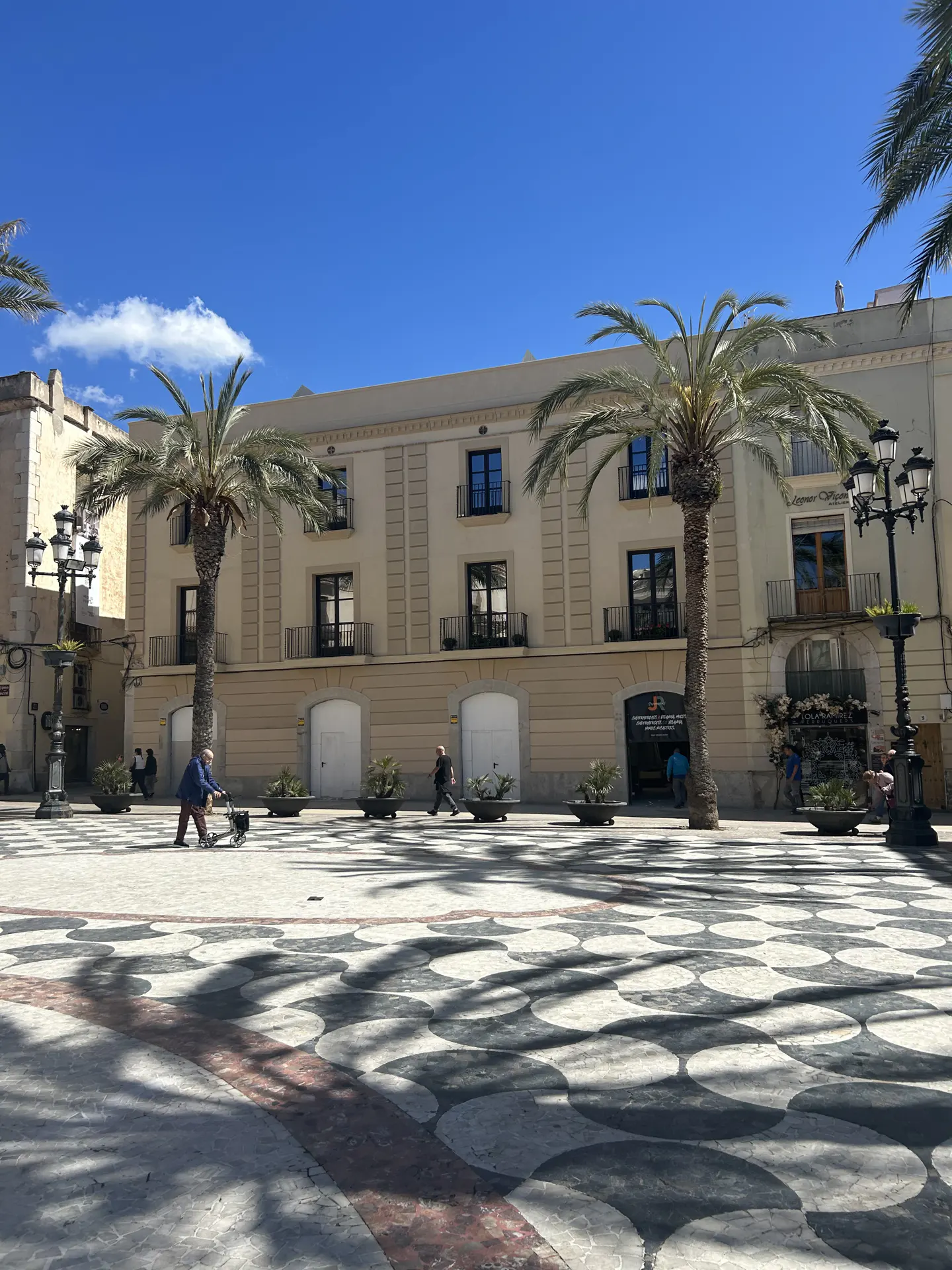 Sunny plaza with palm trees and a beige building. A person uses a walker, and shadows create a patterned ground.