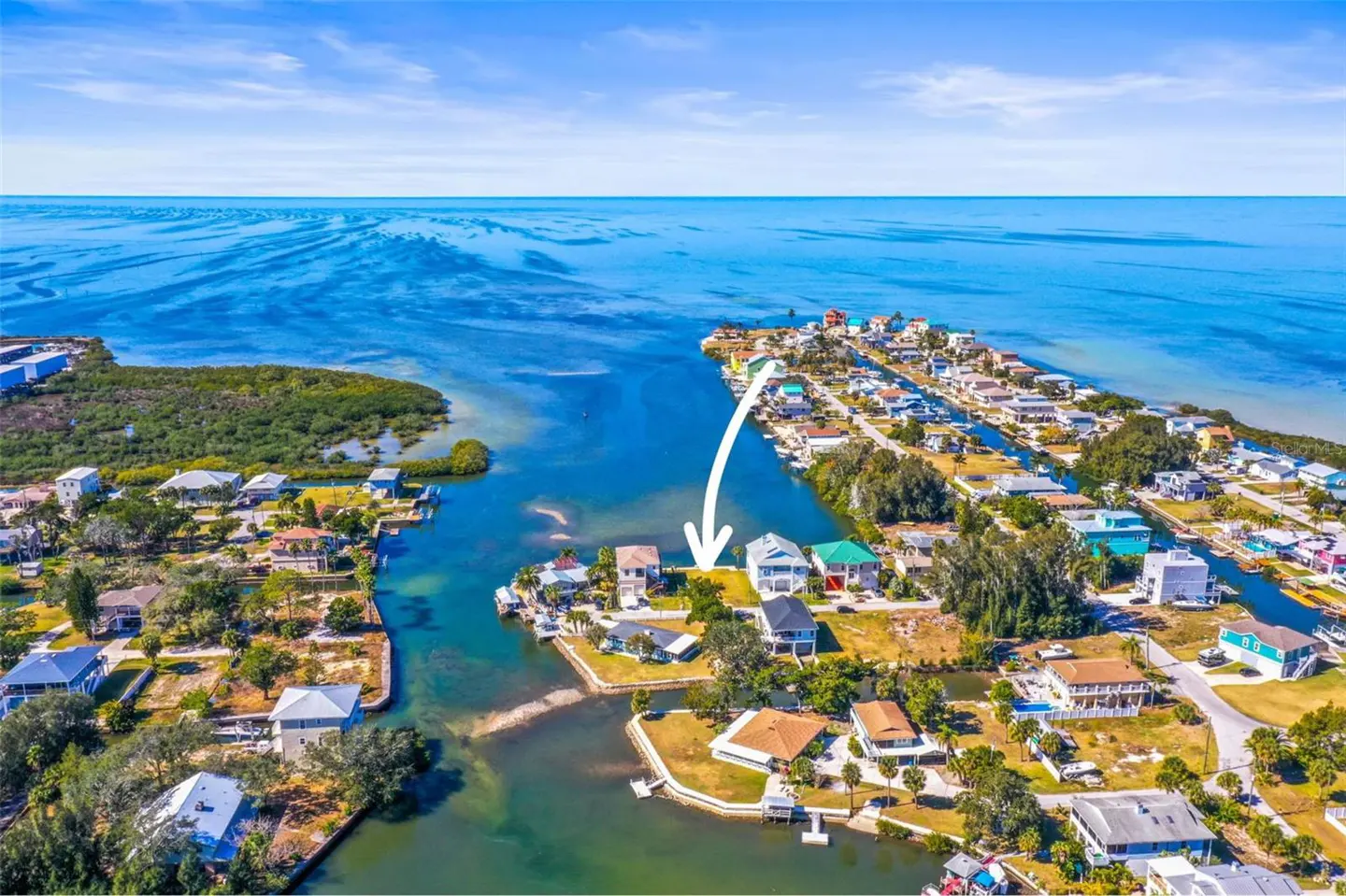 Aerial view of waterfront homes on canals, with an arrow pointing to a vacant lot. Blue sky and ocean in the background.