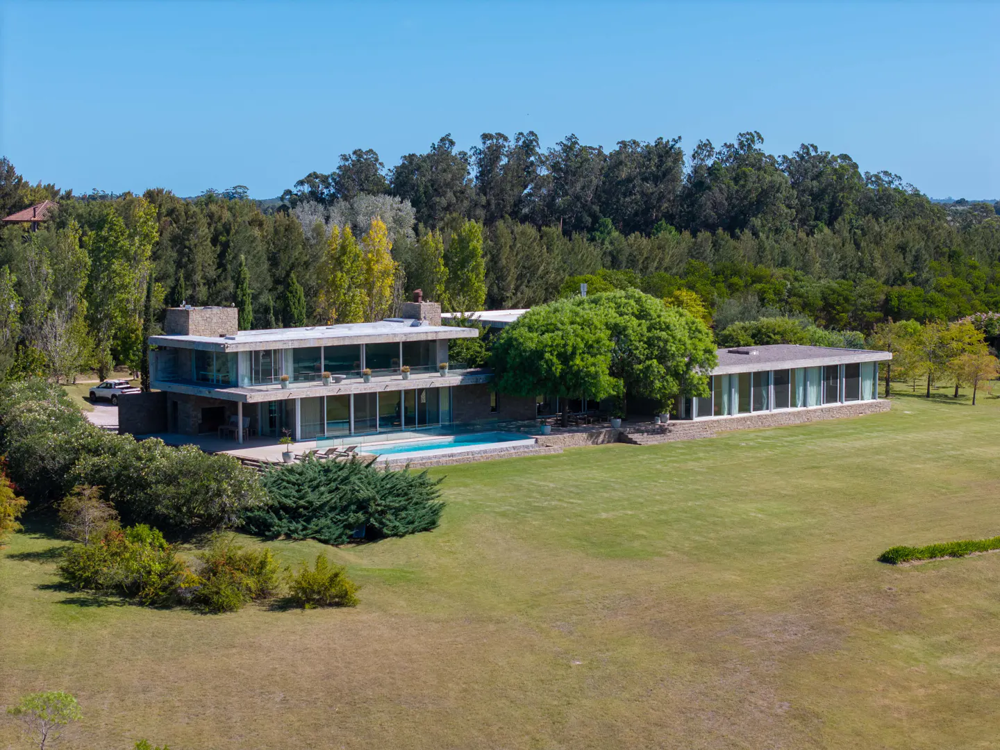 A modern two-story house with a pool, surrounded by green trees and a large lawn.