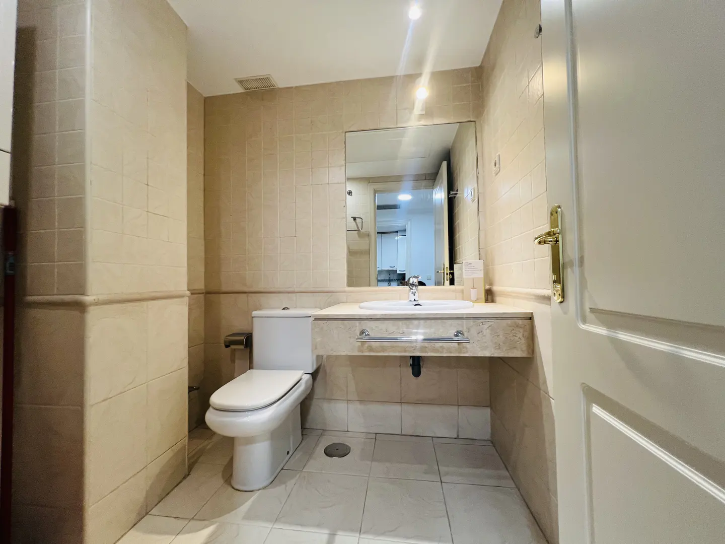 Bathroom with beige tile walls, white toilet, sink with marble countertop, and a mirror. A white door is partially open.
