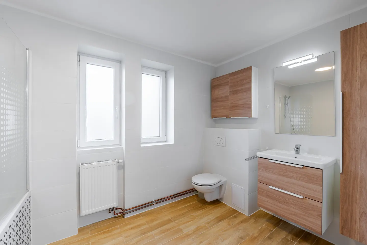 Bright bathroom with wood-look tile, white walls, toilet, sink vanity, mirror, and wood cabinets. Two windows provide natural light.
