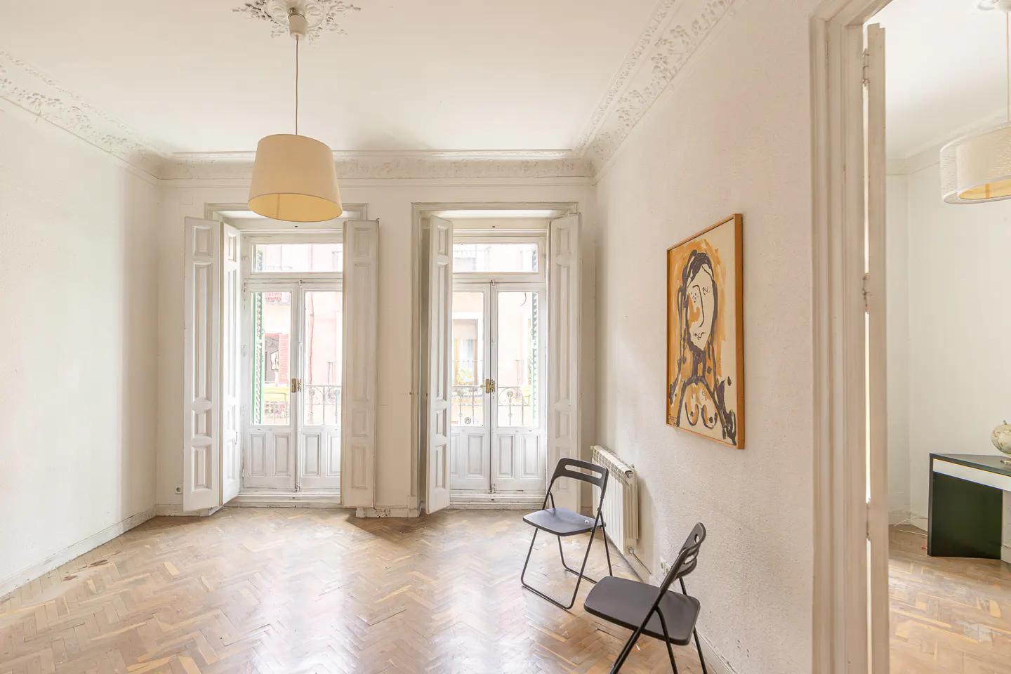 Bright room with herringbone wood floors, white walls, and two sets of white French doors. Two black folding chairs sit near a radiator. A painting hangs on the wall.