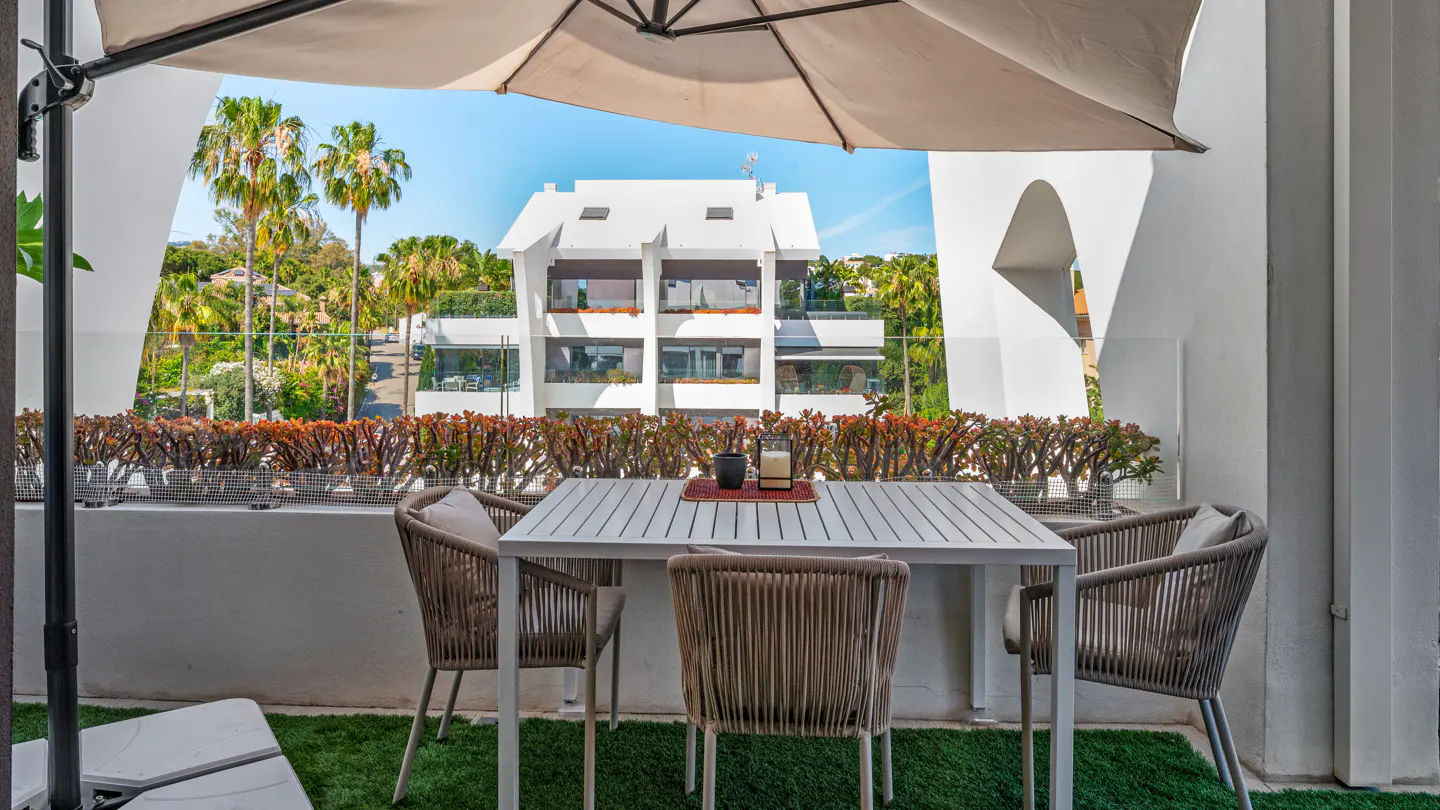 A balcony with a white table and three woven chairs under a beige umbrella, overlooking palm trees and a white building.