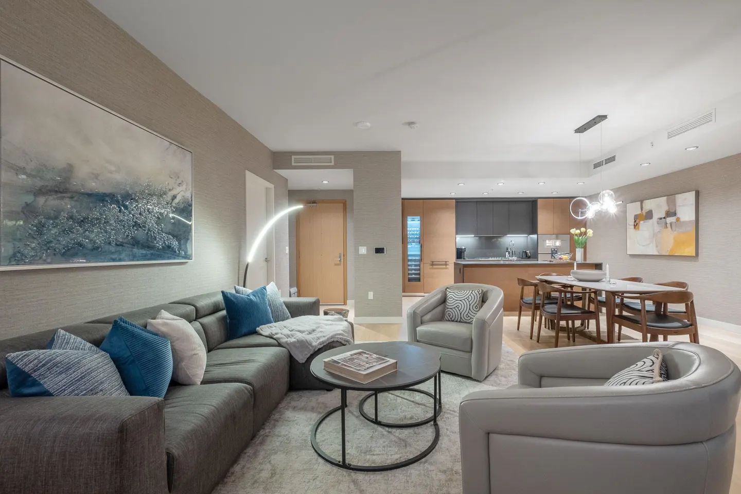 A modern living room with a gray sectional sofa, blue pillows, and a round coffee table. The kitchen and dining area are in the background.